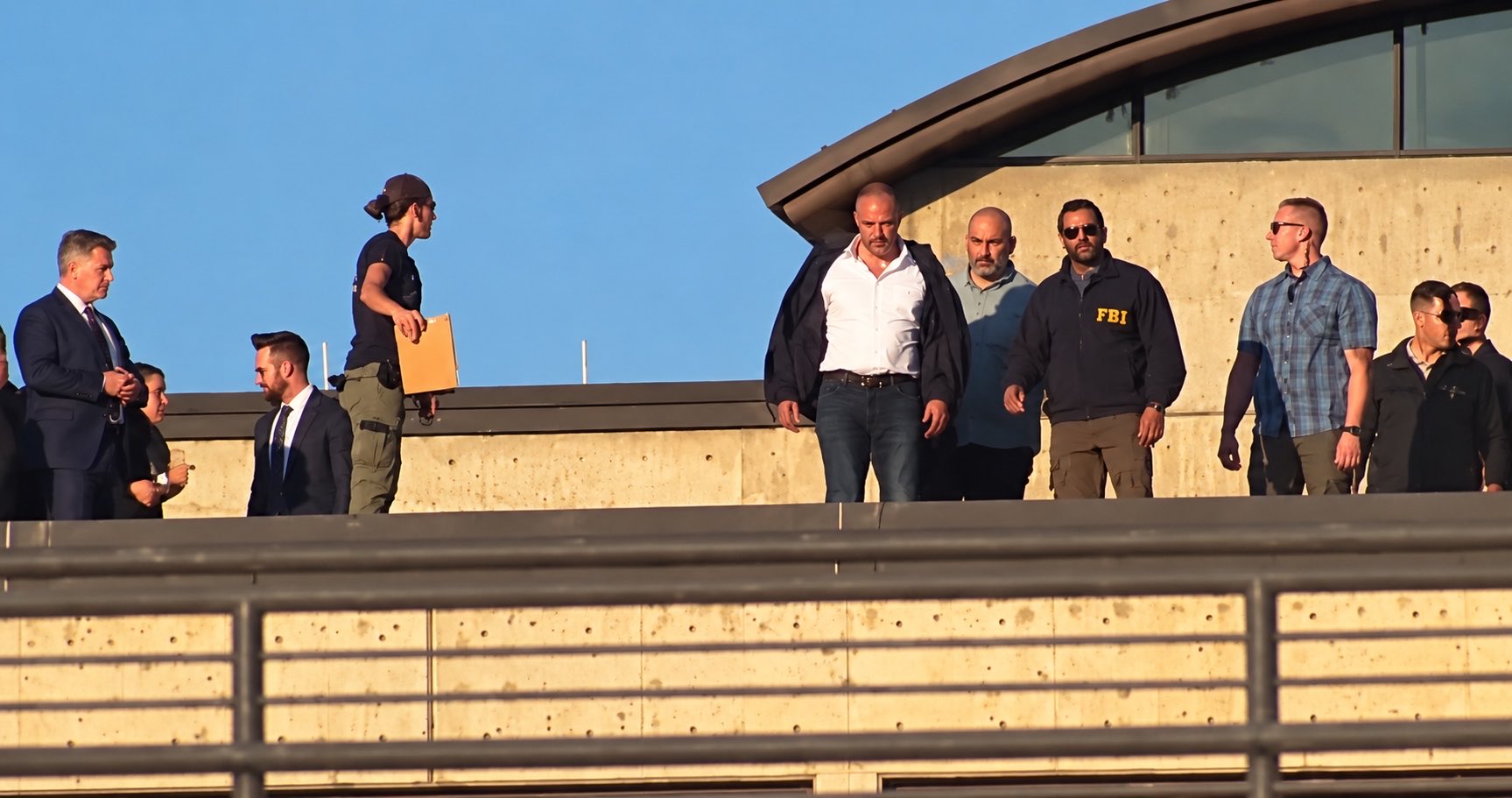 A group of federal and law-enforcement personnel walking a rooftop walkway at Utah Valley University in late-afternoon sun, one man in a navy FBI windbreaker, another carrying a manila envelope.