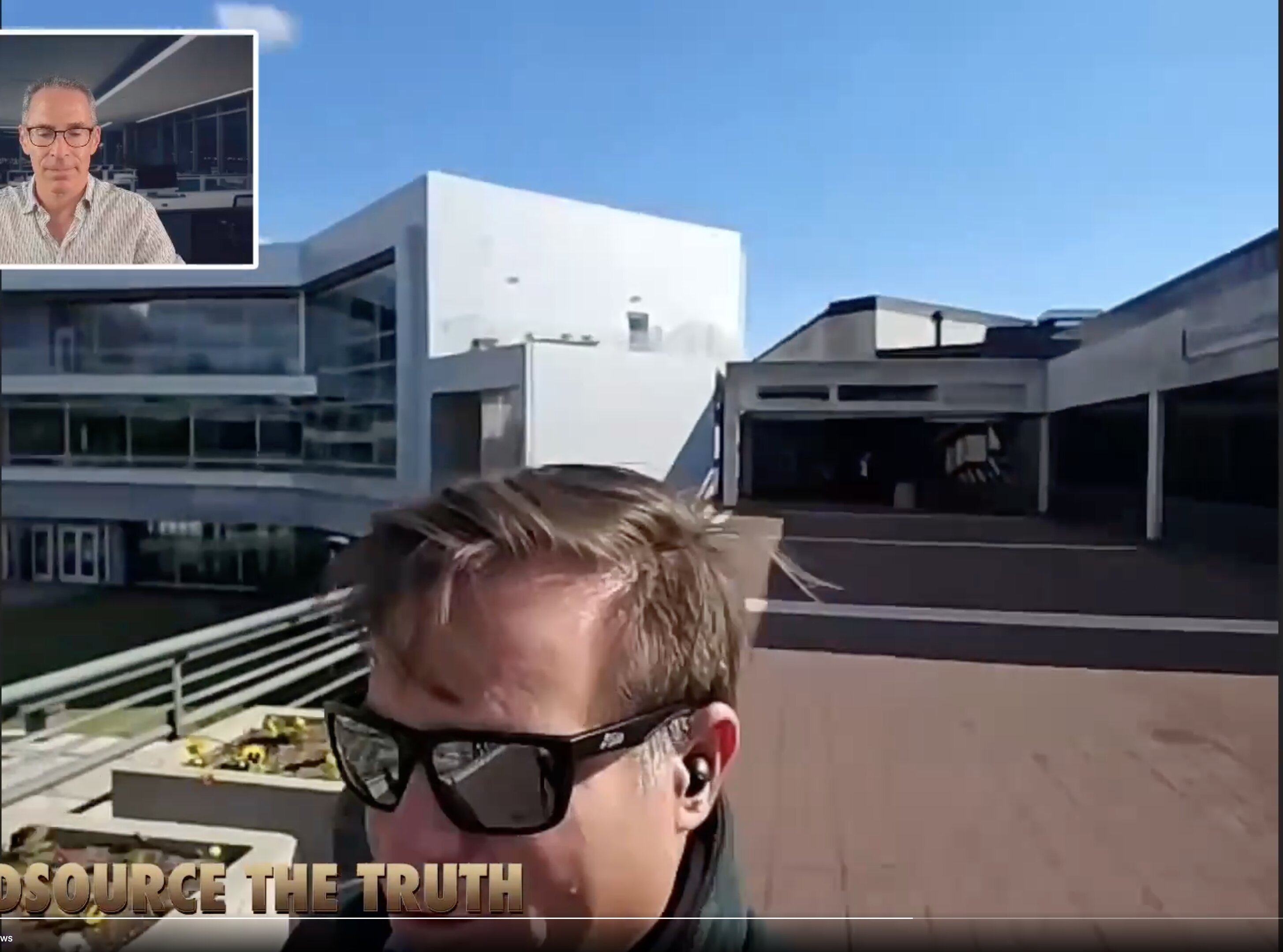 This image is a screenshot from a digital video broadcast or stream, showing a man recording a first-person style selfie video outdoors on a modern paved plaza while another man…