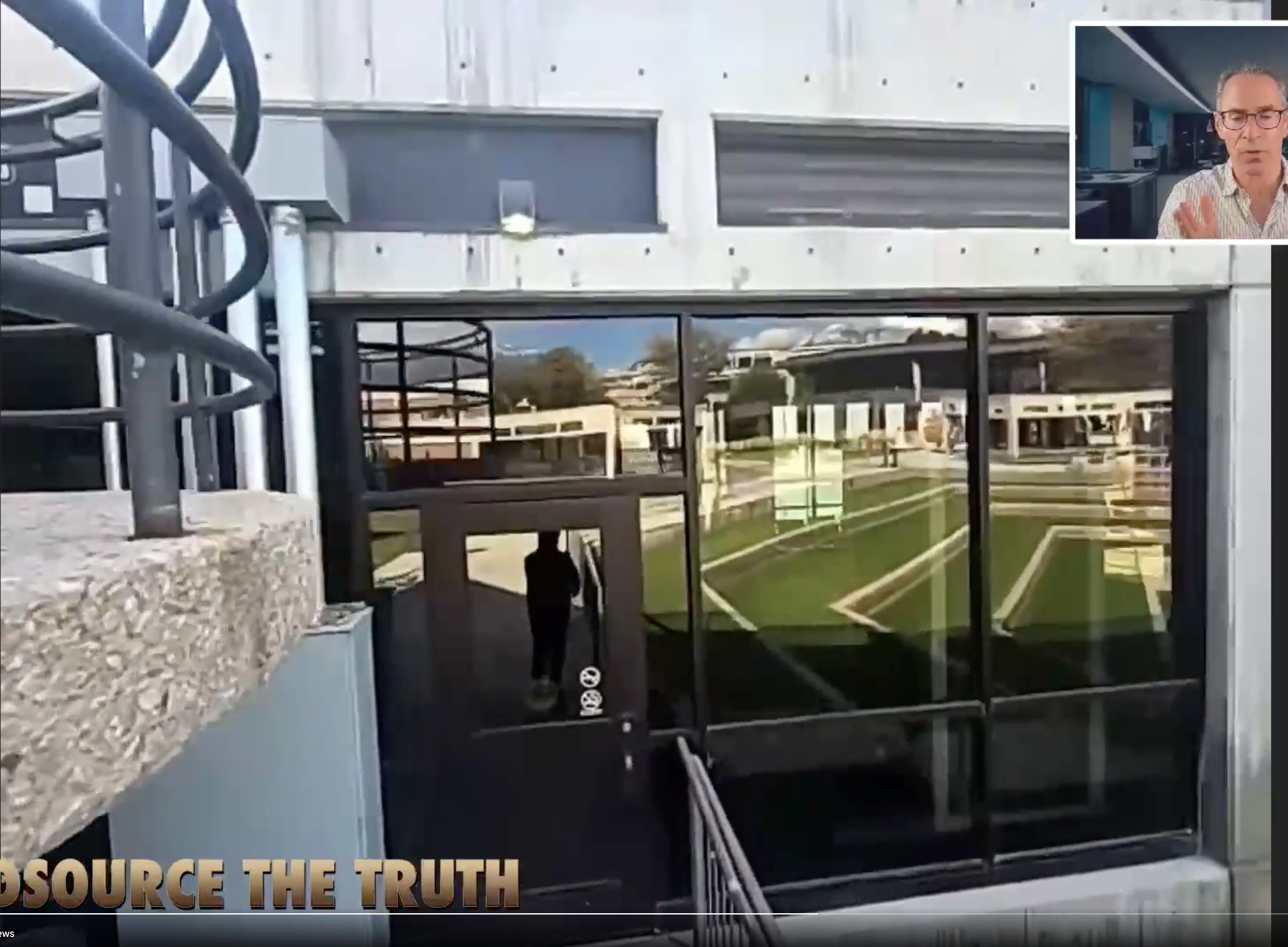 This image is a video frame showing the exterior entrance of a brutalist-style concrete building with large, dark-framed reflective glass windows.