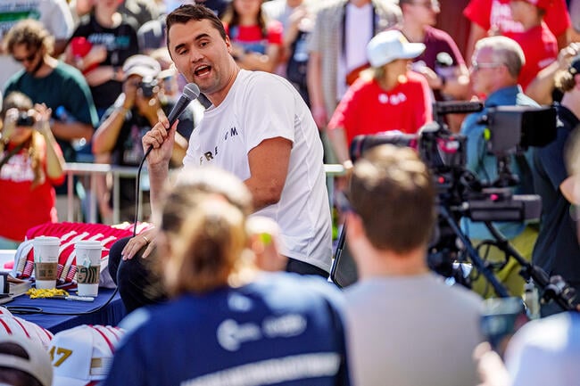 The image shows American conservative activist Charlie Kirk speaking passionately into a microphone at an outdoor public event or political rally.