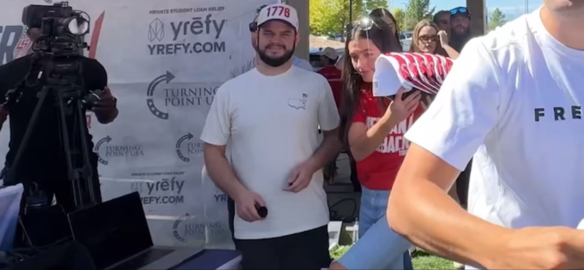 Bearded man in a white Turning Point USA t-shirt and a red-and-white 1776 cap, wearing a headset earpiece, standing in front of a TPUSA and Yrefy step-and-repeat banner, holding a small dark cylindrical object low at his waist.